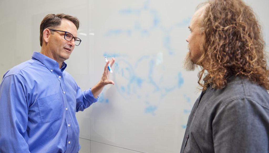 澳门正规博彩十大排行平台 scientists Brett Crawford and Dan Wendt talking by a whiteboard with a scientific formula